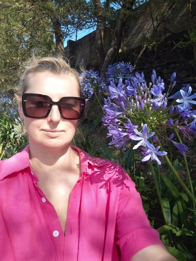 A medium shot features a woman wearing dark-framed sunglasses and a bright pink button-down shirt. She stands outside amidst lush greenery and purple Agapanthus flowers. The location is possibly Ribeira Grande, Portugal, implied by the presence of stone wall behind the flowers, typical of the region's architecture. Trees and foliage provide a background. The sun is shining, casting shadows. The woman faces the camera.