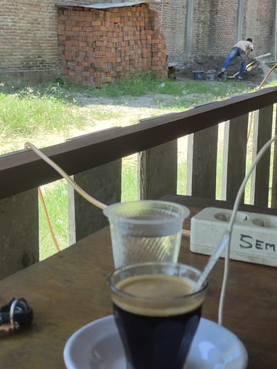 The image captures a scene from Keudematangkuli, Indonesia, focusing on a wooden table in the foreground with a cup of coffee, a glass of water, and a set of keys. In the background, a construction site is visible, featuring a man in blue pants and a light-colored shirt shoveling dirt, with buckets and piles of bricks present. A brick wall and a stack of raw bricks are also prominent in the background. The environment appears to be outdoors, with a wooden railing and concrete posts framing the view of the construction. A white electrical outlet with the word "SEM" written on it is also visible on the table.