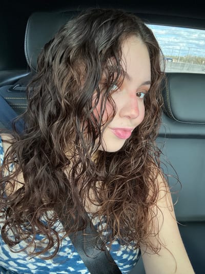 A young woman with long, dark, curly hair and fair skin is positioned in what appears to be a car. She wears a patterned blue and white shirt and a seatbelt is visible across her chest. Her head is tilted to the side, with her gaze slightly upward. The light seems to be coming from the left, casting shadows on her face and hair. The background consists of the car's interior.