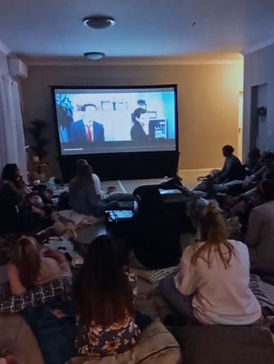The image depicts an indoor gathering in Aktash, Uzbekistan, where approximately 15 individuals, predominantly female, are seated on cushions and blankets on the floor, watching a projection screen. The large screen displays a scene featuring two individuals: a male in a suit and red tie on the left, and a female in dark attire on the right. The room is dimly lit, with ambient light from the screen illuminating the immediate area. Overhead, two recessed ceiling lights are visible. To the left, a white air conditioning unit is mounted on the wall above closed white curtains, with a dark potted plant partially visible below. Towards the right, an open doorway leads to another room or hallway. In the mid-ground, a laptop is open and illuminated, displaying what appears to be data or text. The individuals are mostly facing the screen, their postures indicating passive viewing.