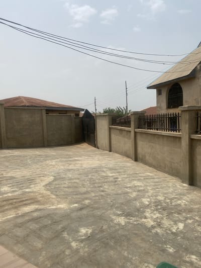 The image depicts an outdoor, paved area within Odo Ona, Nigeria, flanked by unpainted concrete walls. The ground surface is composed of grey concrete pavers with visible wear and discoloration. To the left, a curved, unpainted concrete wall partially encloses a single-story building with a rusted, corrugated metal roof. A dark-colored metal gate, partially ajar, is situated between this wall and a taller, unpainted concrete wall on the right. The right wall is topped with a black wrought-iron fence. Behind this fence and wall stands a multi-story building featuring a light-colored facade, a steeply pitched corrugated metal roof, and a prominent arched window secured with black bars. Overhead, multiple black power lines traverse a hazy, light grey sky with scattered clouds. Utility poles and green foliage are visible in the background beyond the gate and walls. No individuals or active interactions are present.