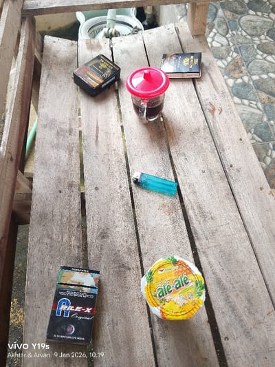A wooden surface displays an assortment of items: two packs of cigarettes (one RILE-X Original, one Sampoerna Kretek), a glass of dark liquid with a red lid, a blue lighter, and a small tub of Ale-Ale pineapple-flavored juice. A passport-sized booklet lies near the glass. The backdrop consists of wooden planks and a section of tiled ground. The lower left corner of the image includes the watermark "Vivo Y19s" and a timestamp indicating "Akhtar & Arvan 9 Jan 2026, 10.19."