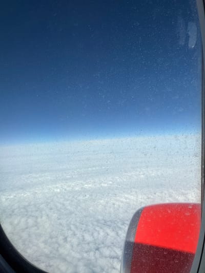 An aerial view from an aircraft window depicts a clear blue sky above a dense, undulating layer of white stratocumulus clouds. The visible aircraft window is heavily speckled with dust or debris. In the lower right quadrant, a portion of a bright red aircraft engine nacelle is visible, also appearing speckled. The scene indicates the aircraft is in flight, soaring above the cloud cover over the region of Granadilla de Abona, Spain. No individuals are visible.