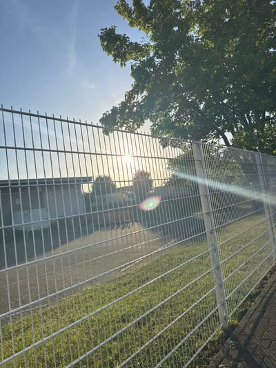 A wire fence runs horizontally across the frame, separating the viewer from a scene bathed in the warm, low light of the sun, which is partially obscured by the dense foliage of a large tree. The fence appears to be a security barrier, likely surrounding an industrial or commercial property. Through the fence, one can see a building with a light-colored facade and what appears to be a white container or trailer parked nearby. The ground in front of the building is paved, and beyond that, a strip of green grass is visible. In the distance, the faint outlines of other structures and trees are discernible. The sky above is a clear blue, with wispy clouds, suggesting it is either early morning or late afternoon. The overall impression is one of a quiet, possibly industrial area on the outskirts of a town, bathed in the golden hues of a setting or rising sun. The image is taken from a slightly elevated perspective, looking across the fence and into the property beyond. There is a slight lens flare visible on the right side of the image, indicating the direction of the sun. Without further information, it is impossible to pinpoint the exact location within Bad Säckingen, Germany.