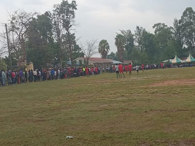 A sports field with a crowd of spectators watching a soccer game. Players in red and white jerseys are spread across the field. A long line of people stands to the left, near trees and a building. A tent can be seen in the background. The sky is overcast.