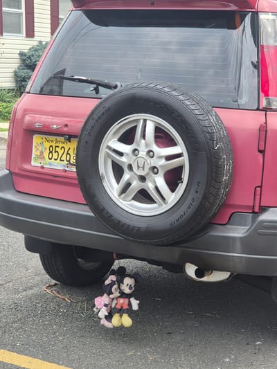 A red Honda CR-V is parked on a paved road with a yellow line. A spare tire is mounted on the back of the SUV. Two plush toys, Mickey Mouse and Minnie Mouse, are hanging from the undercarriage of the vehicle. A New Jersey license plate is visible on the rear of the car. In the background, a house with red shutters and a manicured bush are seen. The scene appears to be in South River, New Jersey, United States of America.
