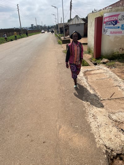 The image depicts a street scene in Barikin Jirga Summa, Nigeria. In the foreground, a person walks on the right side of a paved road. The individual is wearing a dark, wide-brimmed hat, a pink and black jacket over a patterned top, and patterned trousers. They are also wearing light-colored sandals. To the right of the road, there is a concrete drainage channel and a rough, dirt embankment with some vegetation. Behind this, a building with a sign reading "ADMISSION OPEN" and "ELSHAMA INTERNATIONAL SCHOOL" is visible. Further down the street, there are more buildings, including one labeled "LEISURE GAME SHOP." Several utility poles with overhead wires are present along the street, and a few vehicles, including a red car and a motorcycle, are visible in the distance. The sky is overcast.