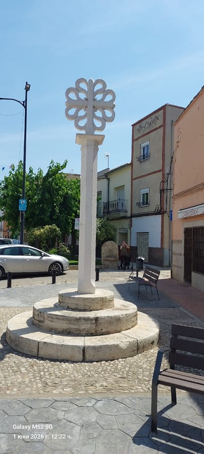 The image shows a roundabout in the city of Canals, Spain. A white stone cross is centered in the roundabout with a decorative, clover-like design at the top. The base of the cross has three circular tiers. A silver car is parked nearby. In the background are several buildings, including one with a sign that reads "CARNISSERIA." An elderly person with a walker is seated on a bench in the background. Other benches and a small, stone structure are also present. The sky is clear and blue. A street lamp with wires extends across the top left of the image. A small blue sign with a white figure is visible near the trees. The pavement is textured, with a combination of smooth, geometric patterns and pebbled sections. The photo includes a watermark with "Galaxy M52 5G" and a date of "1 June 2026."