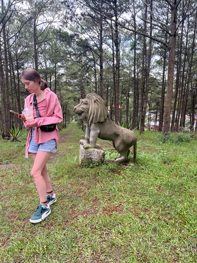 A young woman stands in a grassy area with pine trees in the background. She is wearing a pink button-down shirt over a pink top, blue denim shorts, and green and white sneakers. She holds a clear plastic cup with a straw and a mobile phone in her left hand. A black fanny pack is worn across her chest. To her right, a stone statue of a lion stands on a rock. The environment appears to be a park or garden, possibly located in the Cam Ly area of Da Lat, Vietnam, given the prevalence of pine trees and the style of the stone statue. The ground is covered in green grass and some fallen pine needles.
