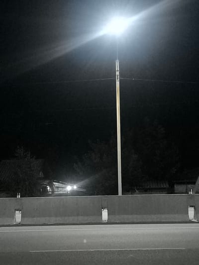 The image depicts a nighttime scene on a road in Belovodskoe, Kyrgyzstan. In the foreground, a concrete barrier separates the road from an embankment. A street light, brightly illuminated, is positioned in the center of the frame, casting a strong light into the dark sky and onto the surrounding area. In the background, beyond the barrier and trees, the silhouette of a building with illuminated windows is visible, suggesting the presence of a residential or commercial area. The overall environment appears to be a rural or semi-urban setting at night. There are no people or animals clearly visible in the image.