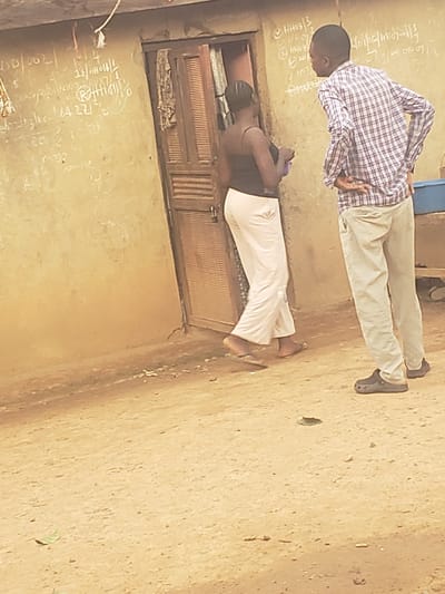 The image depicts two young Black boys in what appears to be an outdoor or semi-outdoor classroom setting. The ground is made of dirt, and there is a rough-hewn wooden structure in the background, possibly a cabinet or doorway with slatted doors. One boy, wearing a checkered shirt and light-colored trousers, is standing and facing away from the camera, with his hands behind his back. The other boy, wearing a dark top and white trousers, is kneeling or crouching in front of a dirt wall, facing towards the same wall where writing is visible.
The wall has chalk writing on it, including numbers, tally marks, and some characters that are difficult to decipher clearly. Some of the visible text appears to be mathematical or educational in nature, with phrases like "16/10/2021", "3/16", and numbers such as "22" and "4". There are also tally marks indicating counts.
The overall scene suggests a casual educational environment, possibly in a rural or less developed area. The lighting suggests it is daytime, and the setting appears to be relatively dry, given the dirt ground. The expressions or emotions of the subjects are not clearly discernible due to their poses and the image quality. The presence of educational writing on the wall strongly indicates a learning activity.