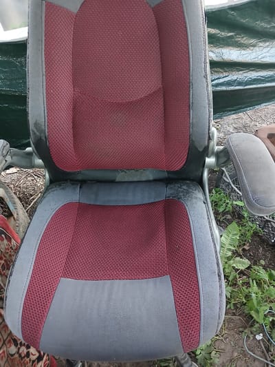 A top-down, medium shot captures a worn, two-toned office chair with burgundy and grey fabric, positioned outdoors in what appears to be a rural or backyard setting in Hlushky, Ukraine. The chair is in disrepair, with visible stains and peeling fabric on the seat and armrests. The background features a green tarp, soil, dried leaves, and some vegetation, suggesting an outdoor storage or discarded item location. No people or animals are present in the frame.