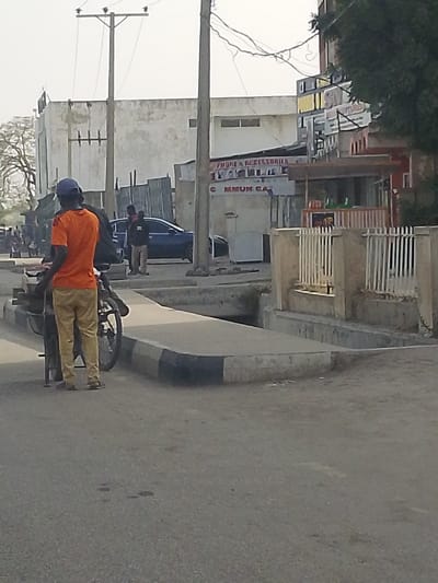 The image depicts a street scene in Gyadi Gyadi Arewa, Nigeria, under bright daylight. In the foreground, a male individual is seen from behind, wearing an orange t-shirt, khaki trousers, dark flip-flops, and a dark blue baseball cap. He stands next to a bicycle fitted with a large, dark metal carrier or cart on its rear wheel. The paved asphalt road occupies the lower portion of the frame.
To the right of the foreground figure, a concrete curb with black and white diagonal markings delineates a raised concrete structure, possibly a sidewalk or drainage system, featuring a visible channel. Further back, a dark blue sedan is parked, and two additional male figures stand near it.
In the mid-to-background, various commercial and architectural elements are visible. On the left, a large, light-colored building with multiple windows stands under a hazy sky. Power poles with numerous overhead wires are prominent. Towards the right, smaller commercial establishments include a shop with a partial sign displaying "PHONE & ACCESSORIES" and "WAKILI" along with "COMMUN CA" (likely "COMMUNICATION"). A reddish-brown awning is visible over another stall. A light-colored concrete wall with white vertical metal railings acts as a fence. Dense green foliage partially frames the right side of the scene.