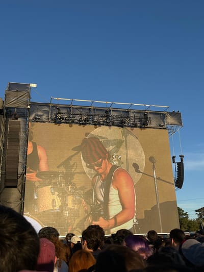 This image depicts an outdoor music concert or festival taking place in San Isidro, Argentina, under a clear blue sky during daylight hours. The central focus is a large LED screen displaying a live feed or recording of a male musician performing. The musician is shown from the chest up, wearing a white tank top, sunglasses, and a red and brown striped beanie, holding an electric guitar. Another musician, partially visible to the left on the screen, is positioned behind a drum kit.
The LED screen is part of a substantial stage structure constructed with metal scaffolding, trusses, and black fabric coverings. Multiple stage lights are visible mounted on the trusses above the screen. Large line array speaker systems are suspended vertically on both the left and right sides of the main screen. The lower portion of the image is filled with the heads and shoulders of a dense crowd, seen primarily from behind, indicating an audience observing the performance. Trees are discernible in the far background, to the right of the stage structure.