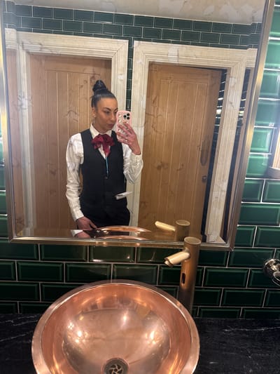 A person in a black vest and white shirt is taking a selfie in a mirror in a bathroom. The bathroom has dark green tiled walls and a copper sink with a brass faucet. The mirror is framed with a marble-like material. The reflection in the mirror shows a wooden door. The location is Warrington, United Kingdom.