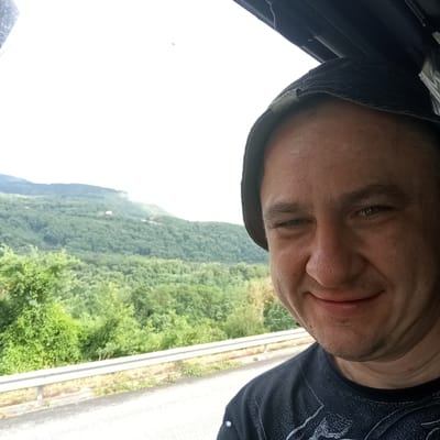 A man wearing a dark patterned t-shirt and a dark baseball cap is taking a selfie. He is positioned on the right side of the frame, with his face taking up most of the right half. The background shows a verdant, tree-covered mountainous landscape, with a road and guardrail visible in the foreground. The sky is bright and overcast. The image appears to have been taken in Saint-Paul-de-Jarrat, France, given the man's attire and the natural scenery.