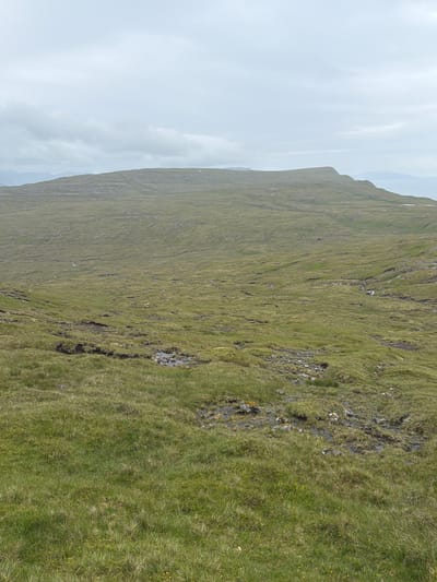 A wide, undulating green landscape under a heavily overcast sky dominates the scene. The terrain is covered in thick, uneven grass with visible patches of darker, damp soil and rocky outcrops. Small streams or rivulets snake through the landscape, indicating a wet environment. In the distance, a broad, flat-topped hill or mountain rises, its edges sharply defined against the muted sky. The overall impression is one of rugged, natural beauty, typical of the Faroe Islands. No people or man-made structures are clearly visible, suggesting a remote, natural setting.