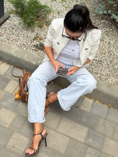 A woman is sitting on the ground, looking at her phone. She is wearing light blue jeans, a white jacket, and a white top decorated with iridescent sequins. Her hair is pulled back in a bun with a black clip, and she is wearing black-framed glasses. Her feet are adorned with strappy leopard-print high heels. A brown handbag with a gold zipper and a small teddy bear charm is placed next to her. The setting appears to be outdoors, with a mix of grey gravel and paving stones. A small evergreen bush is visible to her left. The lighting suggests it is daytime. The woman's posture and focus on her phone indicate she is engaged with its content.