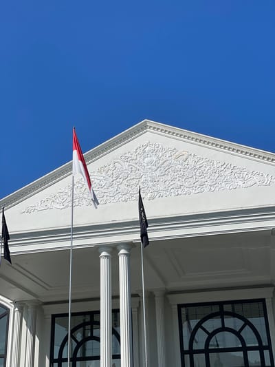The image displays the upper portion of a white neoclassical-style building against a clear, vibrant blue sky. The sky occupies the upper half of the frame, devoid of clouds. The building's prominent feature is a white triangular pediment adorned with intricate white bas-relief floral and acanthus scrollwork patterns. A subtle, light gold-colored accent is visible within the relief on the left side of the pediment.

Three flags are visible:
1.  Centered on the facade is a red and white bicolour flag, consistent with the national flag of Indonesia, attached to a silver flagpole and hanging vertically.
2.  To the left of the Indonesian flag, a black flag with partially visible white or light-colored text (appearing as "A A A") is mounted.
3.  To the right of the Indonesian flag, another similar black flag with white or light-colored text is also mounted.

Below the pediment, two white fluted columns are visible, supporting an overhang. The underside of this overhang features a white paneled ceiling. At the bottom of the frame, portions of large dark-framed windows or glass doors with arched tops are visible, reflecting ambient light. No individuals are present in the scene. The setting is in Kayuambon, Indonesia.