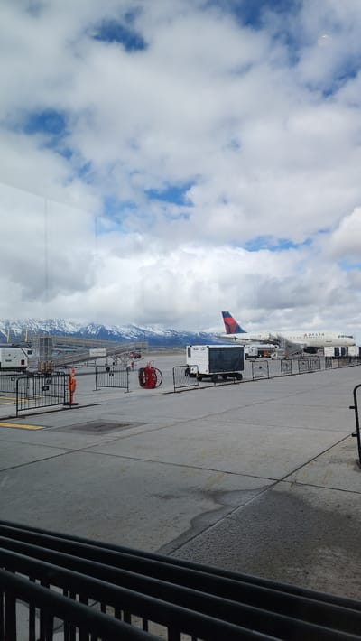 This image captures a view from inside a building, likely an airport terminal, at Jackson Hole Airport in Jackson, United States. Through the window, a Delta Airlines plane is visible parked on the tarmac. Ground crew equipment, including a baggage vehicle and a fire hose reel, surrounds the aircraft. In the background, mountains, partially covered in snow, are visible under a sky filled with clouds and patches of blue. The scene is mostly static with no clear actions or interactions visible.