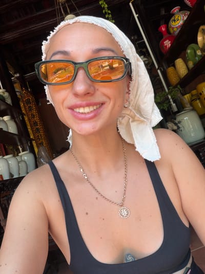 A bright, close-up selfie captures a person with a radiant smile, looking directly at the camera. The individual wears a light-colored, textured head wrap and black-framed sunglasses with orange-tinted lenses, which reflect the indoor environment. A delicate gold-colored chain necklace with a prominent circular pendant featuring a sparkling rim and an intricate central design adorns their neck. They are wearing a black sleeveless top, and a spiral-shaped tattoo is visible on their upper chest/shoulder area. Earrings are visible on the person's right ear (viewer's left).
The background suggests an indoor setting, possibly a craft shop or traditional retail space in Lương Sơn, Vietnam. Wooden shelves are filled with a diverse collection of pottery and ceramic vessels. These items display a variety of colors, including red, green, yellow, and white, with many featuring hand-painted floral and decorative motifs. Some pieces, such as a red vase in the upper left, have text that appears to be stylized Vietnamese script. Green plants are also integrated into the display. Behind the person's head, a golden decorative panel or scroll with dark, ornate Vietnamese or Chinese characters is clearly visible. Additional stacks of white ceramic items are seen in the lower right portion of the image. The bright lighting indicates it is daytime, and the person's broad smile conveys a sense of happiness.