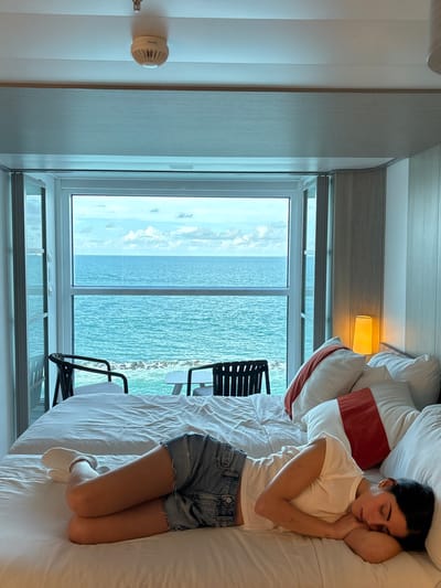 A female individual is resting on her left side on a large white bed in a contemporary room. She is wearing a white short-sleeved top, light blue denim shorts, and white socks, with her eyes closed, indicating sleep. The bed has white linens and multiple pillows, two of which display a reddish-orange accent band. To the right of the bed, a minimalist table lamp provides warm illumination. A prominent window offers a direct view of the Atlantic Ocean, displaying turquoise water under a partially cloudy sky. Outside the window, two dark-colored outdoor chairs and a small white table are visible on an exterior balcony or terrace. Below the window, concrete coastal defense tetrapods are present in the water, indicating proximity to the shoreline. A white smoke detector is mounted on the ceiling. This scene depicts an interior space within a beachfront high-rise property in Miami Beach, Florida, United States.