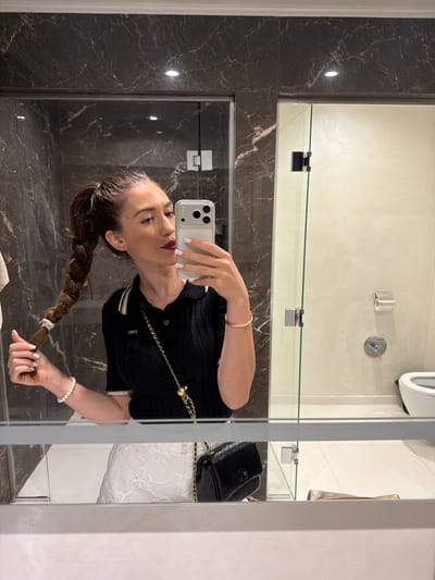 This is a mirrored selfie taken in a bathroom. The subject is a woman with long, braided brown hair, wearing a black top with a white collar and trim, and a pearl bracelet on her left wrist. She is holding a smartphone in her right hand, which is capturing the reflection. In the reflection, a black handbag with a gold chain strap is visible. The bathroom features dark marble walls and a large mirror. The woman appears to be taking a photo of herself while looking at her reflection. The overall mood is casual and personal.