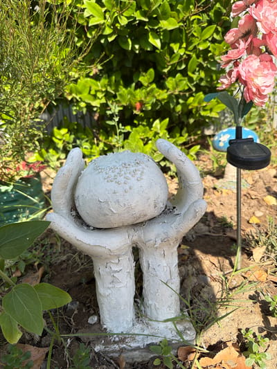 The image depicts a white, textured garden sculpture of cupped hands holding a round object, likely a decorative ball, positioned in the foreground. The sculpture appears aged, with peeling paint. Behind it, lush green foliage creates a natural backdrop. To the right, a solar-powered garden light with a black top is visible, mounted on a thin metal stake. Nearby, a blue mushroom-shaped ornament adds a touch of whimsy. The ground is a mixture of soil, fallen leaves, and sparse green vegetation, suggesting an outdoor, possibly garden or yard setting. The sunlight casts shadows, indicating a daytime scene. The image does not contain any discernable elements that would specifically place it within Balakənd, Azerbaijan; it depicts a generic outdoor scene.