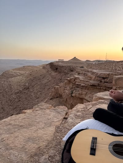 The image depicts a serene moment at a cliff edge in Mitzpe Ramon, Israel, during either sunrise or sunset. The sky presents a striking gradient, transitioning from a soft, cool grey-blue on the left to warm, vibrant orange and yellow tones on the right, indicating a clear, calm atmosphere.
In the foreground, on the right, a person is partially visible, seated on the rugged, rocky ground. They are wearing black pants, and their hands are subtly clasped. Resting beside them is a light-colored classical acoustic guitar, partially enclosed in a black gig bag with a white cover. The guitar's soundhole, black bridge, and white strings are clearly discernible.
The setting is a dramatic, arid landscape viewed from atop a high cliff. The foreground features weathered, stratified rock formations, characteristic of a desert environment. Beyond the immediate cliff face, a vast expanse of barren, rocky desert stretches into the distance, with distant hills and plateaus under the expansive sky. Along the far rim of the cliff, faint structures, possibly buildings or communication towers, can be observed. The scene evokes a sense of peace and contemplation, with the individual seemingly enjoying the panoramic views and quiet solitude with their instrument. There is no visible text.