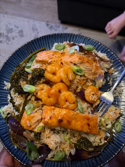 A plate of food is viewed from above. The food appears to consist of rice, pieces of salmon, shrimp, and greens, with a fork placed on top. The plate has a dark blue, ridged surface. A partial view of a person's foot is visible in the upper left corner of the image, indicating the food is likely being held by a person. The background is slightly blurred. The location details are not discernible.