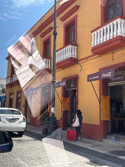 Here is a concise description of the image:
The image depicts a street scene in Veracruz City, Mexico, in front of a restaurant called "La Carreta". The restaurant has a yellow and red façade with white balustrades. Coca-Cola banners are displayed above the restaurant's entrance. A menu board lists various seafood dishes. On the sidewalk, a person walks with a red shopping bag and a white tote bag. A white Nissan Sentra is parked nearby. The image is taken from inside a vehicle, as evidenced by the reflection on the windshield.