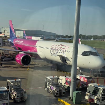 The image shows a Wizz Air airplane parked at an airport gate. The airplane is white with large pink and blue "WIZZ" lettering on its fuselage. The Wizz Air logo is also visible on the engine. Several airport service vehicles are parked around the plane, including a baggage tractor and a tow tractor. One of the tow tractors has the text "London Luton Airport" visible. The scene appears to be an active airport tarmac on a sunny day, with clear blue skies. The image is taken from a slightly elevated position, possibly from an airport terminal window, looking down at the plane and ground operations. The overall atmosphere is one of routine airport activity. The text "wizzair.com" is visible multiple times on the aircraft.