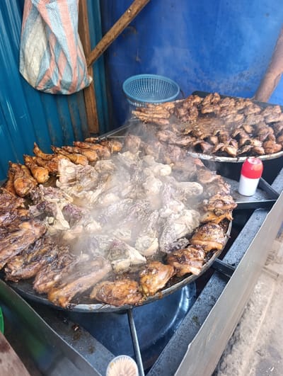 This vivid image captures an outdoor street food stall in Kahawa Wendani ward, Kenya, during daylight hours. The primary focus is on two large, round metal griddles or pans filled with numerous pieces of meat, predominantly chicken, in various stages of preparation.
In the foreground, a griddle holds a substantial quantity of chicken pieces, including drumsticks, thighs, and smaller cuts. Visible steam rises from the cooking meat, indicating that it is actively being cooked or kept warm over heat. Some pieces are dark brown and appear well-cooked and caramelized, while others are lighter in color, possibly having just been added or still requiring more cooking time. The meat seems to be simmering in its juices or a marinade. To the right, in the background, a second, slightly smaller griddle is also packed with cooked or nearly cooked meat.
The cooking apparatus consists of sturdy metal frames supporting the griddles. Adjacent to the background griddle, a red and white plastic dispenser bottle, likely containing oil, water, or sauce, is placed on the metal stand.
The background features blue corrugated metal sheets, forming a wall or enclosure for the stall. A light blue plastic basket hangs on this wall. In the upper left corner, a large, striped bag (with white, blue, and reddish-pink stripes) hangs from a rustic wooden beam. More wooden planks are visible as part of the makeshift structure. The scene portrays a bustling and practical outdoor kitchen, preparing a large volume of meat for sale. There is no discernible text in the image.