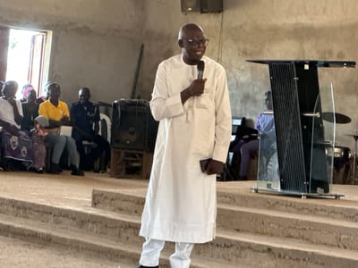 In Kufana, Nigeria, a Black man wearing a white kaftan and trousers stands on concrete stairs in front of a podium, speaking into a microphone. He is wearing glasses and has a bald head. Behind him, other Black individuals are seated, some wearing traditional attire. A large speaker system is visible to the left, and a window with natural light is on the far left wall. The environment appears to be an indoor gathering space, possibly a church or community hall.