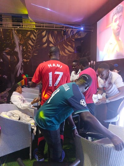 The image depicts a scene in a possible social event in Syokimau-Mulolongo ward, Kenya. A woman with braids is seated in a wicker chair. A man wearing a red jersey with "NANI 17" on the back stands nearby. Another man, in a teal jersey with "ROSSIA" on the back, moves wicker chairs. Two men in pink and white shirts stand together, one is using paper towels. On a screen in the background, a man is shown. Light beams emit from above. Balloons in Kenyan flag colors are present.