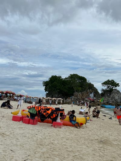 This image depicts a crowded beach scene at Phru Nai, Thailand, on a cloudy day. In the foreground, a yellow and orange inflatable banana boat is positioned on the sand, laden with numerous orange and green life vests. Several red rectangular buoys are visible supporting the boat.
People are scattered across the sandy beach. Some are seated on the sand, while others stand or walk. In the middle ground, a cluster of beach umbrellas in various colors shades a group of people. Further back, rocky outcrops covered with lush green trees rise from the sea. Some individuals are seen near these rock formations.
The ocean is visible in the background, with gentle waves lapping the shore. The sky is overcast with a mix of light and dark clouds. Several other inflatable water toys, including a blue floating mat and a green kayak, are present near the water's edge. A few individuals are dressed in swimwear, and one man in the lower right corner wears bright orange swim trunks. The overall impression is a busy recreational beach setting.