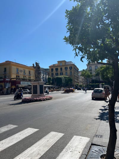 A statue of a religious figure, likely Saint Anthony, stands on a pedestal in the middle of a street intersection in Sorrento, Italy. The statue is surrounded by a circular planter filled with pink flowers. In the foreground, a crosswalk is visible on the asphalt road. Several vehicles, including scooters and cars, are present in the intersection and on the surrounding streets. Buildings with multiple stories line the sides of the street, some with balconies and awnings. Trees, including one in the foreground to the right, provide shade. The sky is clear and blue, indicating a sunny day. People are seen walking on the sidewalks and riding on the scooters. The overall scene depicts a bustling public square in an Italian city.