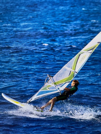 A windsurfer rides a board on the ocean. The windsurfer is dressed in black and green. The sail has white, green, yellow, and blue design elements. Water sprays off of the side of the board. The ocean appears to be deep blue. The location is not evident from the image.