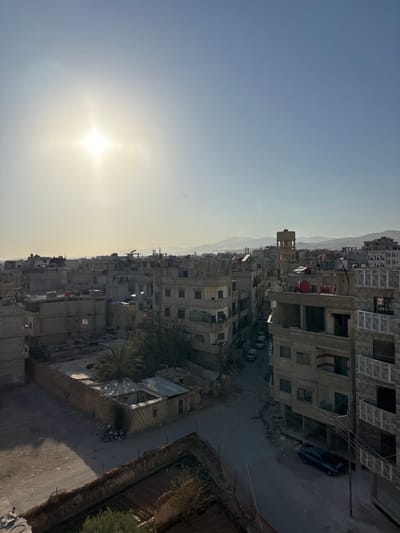 This image captures an elevated view of a residential area in Mu'adammiyat ash-Sham, Syria, under clear daylight conditions with a low sun angle. The sun is prominently featured in the upper-left quadrant, emitting a bright flare.
The scene is dominated by a dense arrangement of multi-story buildings, predominantly constructed from light-colored concrete or block materials. Many structures appear utilitarian, some exhibiting exposed rebar or unfinished sections, while others feature flat rooftops equipped with red cylindrical water tanks. Corrugated metal roofing is visible on lower annexes or damaged sections of some buildings in the foreground.
Narrow, paved and unpaved streets wind through the architectural landscape. A dark blue sedan is parked on a street in the lower-right portion of the frame. Two additional lighter-colored sedans are partially visible further down a street in the mid-ground, leading toward the center-left.
A distinctive, tall, cylindrical water tower-like structure with a square base stands in the mid-ground, slightly to the right of the image center. In the lower-middle left, a low-rise enclosed structure with a partially de-roofed or collapsed section is visible, surrounded by accumulated debris. An empty, walled, rectangular enclosure with exposed earth occupies the immediate foreground, bottom center.
Vegetation is sparse, primarily consisting of several palm trees within enclosed courtyards and scattered smaller green foliage. An intricate network of overhead power or communication lines crisscrosses between buildings and utility poles. No individuals are visible, and no overt actions or interactions are depicted. Distant, hazy mountain ranges are discernible along the horizon.