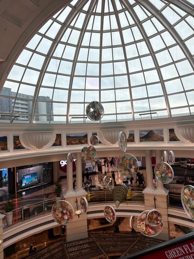 The image shows the interior of a building, potentially a shopping mall, in Milnerton, South Africa. The most prominent feature is a large, curved glass roof supported by a white grid structure. Hanging from the ceiling are decorative floral arrangements encased in transparent discs. Below, there are multiple levels of shops and walkways, with people walking around. There are also some architectural details like pillars, arches and paintings on the wall. Outdoor buildings are visible through the glass roof. There are digital displays showing advertisement of "ConnectLife", "Win" and "100" on one side of the hall.