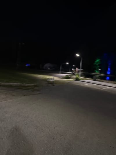 The image shows a dark, outdoor scene, likely at night in the city of Turgoyak, Russia. A paved area, possibly a parking lot or road, dominates the foreground, with a slight incline and visible texture. A grassy area is visible to the left, with a low concrete barrier along its edge. In the mid-ground, a lamppost with a bright light illuminates the scene. Further back, there are more lampposts, some with visible lights, and a structure that could be a building or a fence. In the background, a body of water reflects colorful lights, appearing green and blue, suggesting artificial illumination. The overall impression is one of an urban or semi-urban environment at night, with artificial lighting being a prominent feature.