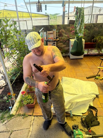 The image depicts a man in an outdoor greenhouse or sunroom environment. The man, appearing shirtless and wearing a checkered cap and camouflage pants, is holding a metal pipe in his left hand and a green bottle in his right. He appears to be in the process of working on something with the pipe and bottle. The environment is cluttered with various objects including a large white bag, a green hanging cushion, plants in pots, a toy kitchen set, and gardening tools. The floor is tiled, and the structure appears to be made of metal and plastic sheeting. The background shows greenery and a distant landscape under a partly cloudy sky. No specific city or location like Valdgeym, Russia is visible or suggested in the image.