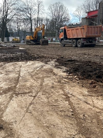 The image depicts a construction site in Donchevo, Bulgaria, under an overcast sky. The foreground consists of heavily disturbed earth, showing a mix of dry, cracked soil and darker, wet mud, with clear tire and track impressions from heavy machinery. In the midground, a yellow Hyundai crawler excavator is parked on the left, its bucket resting on the ground. Further left and behind the excavator, a smaller yellow vibratory roller is also present. To the right, an orange multi-axle dump truck (e.g., MAN TGS) is parked facing away from the viewer, its bed empty. The background features a line of leafless deciduous trees, suggesting late autumn or winter. Several utilitarian structures are visible behind the machinery, including a white construction fence, a small grey building with a red roof, and industrial-like components. No personnel are visible.