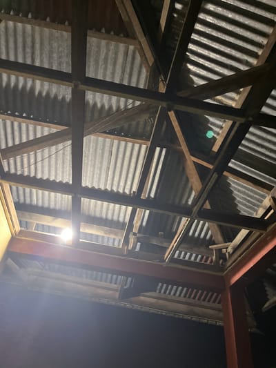 This media file shows the underside of a roof structure, likely at night, given the dark sky. The roof is constructed with a combination of corrugated metal sheeting and wooden beams. A red steel beam is also visible, suggesting a mixed-material construction. The scene is illuminated by a bright, white light source, possibly a bulb, which casts strong shadows on the corrugated metal.

The wooden beams form a complex, intersecting framework, supporting the metal roofing. The corrugated texture of the metal is clearly visible, creating a ribbed pattern. The overall impression is of an unfinished or utilitarian structure, perhaps a barn, shed, or an open-sided building. There are no people visible in the frame.

The context provided is "Kota Kuala Simpang, Indonesia." While the image itself doesn't offer specific visual cues to pinpoint this exact location, it depicts a common type of construction seen in many tropical regions. The time of day is clearly night, indicated by the dark sky and the artificial lighting. No specific weather conditions are apparent. The overall mood is functional and perhaps a little stark due to the lighting and construction materials. There is no visible text in the image.
