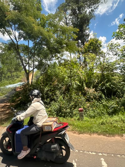 A person rides a scooter on a dirt road lined with lush green foliage and trees under a bright blue sky with scattered clouds. The rider is wearing a light-colored jacket, dark pants, a white helmet with black lettering, and white Crocs. A blue bin is attached to the front of the scooter, and a small cardboard box is secured to the rear rack. The environment suggests a rural or semi-rural setting within Thôn 6, Vietnam, characterized by dense vegetation and a natural landscape. A red and white marker post with the inscription "H8 110" is visible on the right side of the road, indicating a location marker. The road itself appears to be unpaved, with patches of sunlight and shadow.