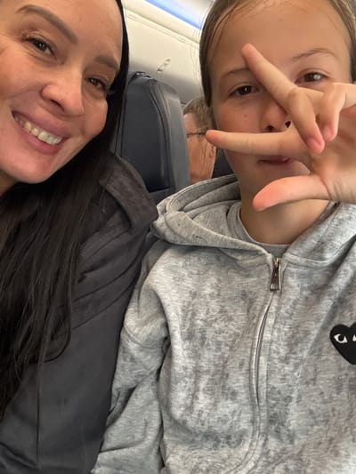The image shows a close-up selfie taken inside an airplane cabin, as indicated by the overhead compartment and airplane seating visible in the background. Two individuals are in the frame. On the left, a woman with dark hair is smiling at the camera. On the right, a child is making a peace sign gesture with their fingers in front of their face. Both are wearing casual clothing. There is no indication of the city of Shirkovo, Russia in the image.
