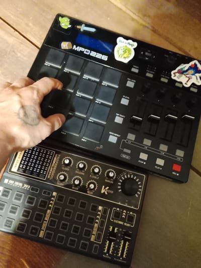 A hand with a tattoo is pressing a pad on a black MPD226 drum machine, which is adorned with cartoon stickers. The drum machine sits atop another black electronic device with multiple knobs, buttons, and sliders. The scene is situated on a wooden surface. There is no information in the image to establish location in Высокиничи, Russia.