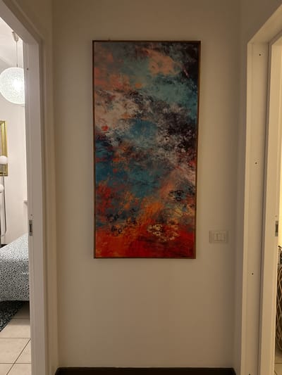 The image shows a horizontally-oriented abstract painting with a bronze-colored frame mounted on a white wall above a doorway. The painting features a blend of colors including shades of blue, black, white, and orange. Above the painting is a light switch with three buttons. The doorway reveals a glimpse into another room with a patterned armchair, a spherical chandelier, a metal clothing rack with white spheres, and white tile flooring. The location is not discernible from the image and cannot be narrowed down to Perugia, Italy, or any specific geographic location.