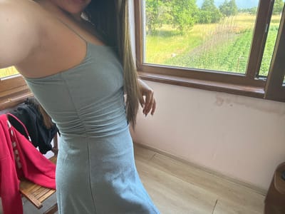 A person is shown from the waist down, wearing a shimmering silver dress. Their hand, with manicured nails painted blue, rests on their hip. Long, light brown hair cascades down their back. The setting appears to be indoors, near a window that looks out onto a lush green landscape with trees and a garden. Sunlight streams through the window, suggesting it's daytime. The scene is casual, possibly a candid moment captured in a home. The overall mood is relaxed and perhaps a bit playful.