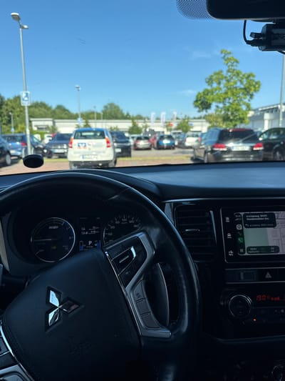 The image is taken from inside a car, looking out through the windshield and at the car's dashboard. The dashboard is equipped with a navigation screen displaying a map and directions, a temperature gauge reading 19.0 degrees, and various controls. A steering wheel with a Mitsubishi logo is prominent in the foreground.
Outside the car, a tree with green foliage is visible against a clear blue sky, suggesting a sunny day. A row of parked cars and what appear to be lampposts line a street or parking lot. The navigation screen indicates a destination on "Hundsmühler Str." in Oldenburg, Germany, and shows the time as 19:01. The overall scene depicts a parked car, possibly in a parking lot or on a street in Oldenburg, Germany, on a pleasant, sunny day. The view through the windshield suggests the car is stationary.